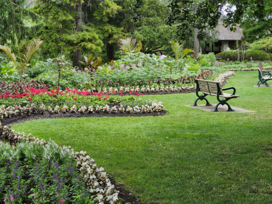 A well-maintained public garden features lush, neatly trimmed green grass with a winding border of colorful flowerbeds filled with pink, white, purple, and green foliage. Tall trees with dense green l
