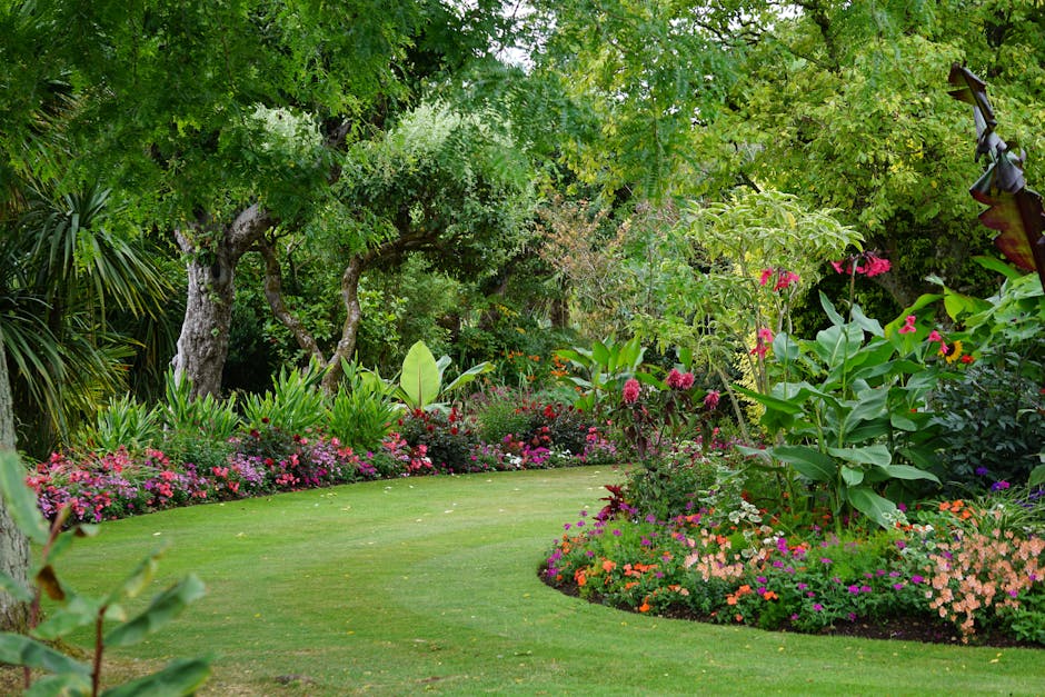 A well-maintained public garden features lush, neatly trimmed green grass with a winding border of colorful flowerbeds filled with pink, white, purple, and green foliage. Tall trees with dense green l